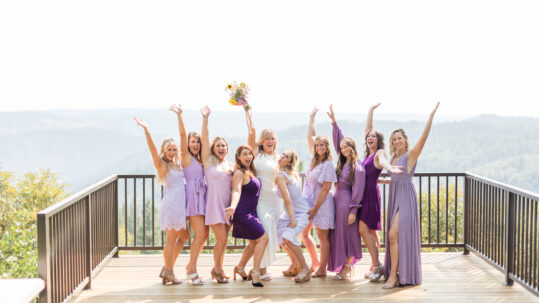 A bride and bridesmaids in purple dresses standing on a deck in front of a mountain background.